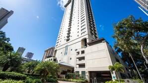 Un immeuble moderne et imposant, doté de nombreux étages et de balcons, entouré d’une végétation luxuriante et d’autres bâtiments de grande hauteur.