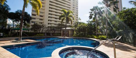 A swimming pool with a hot tub, surrounded by palm trees and a tall building in the background.