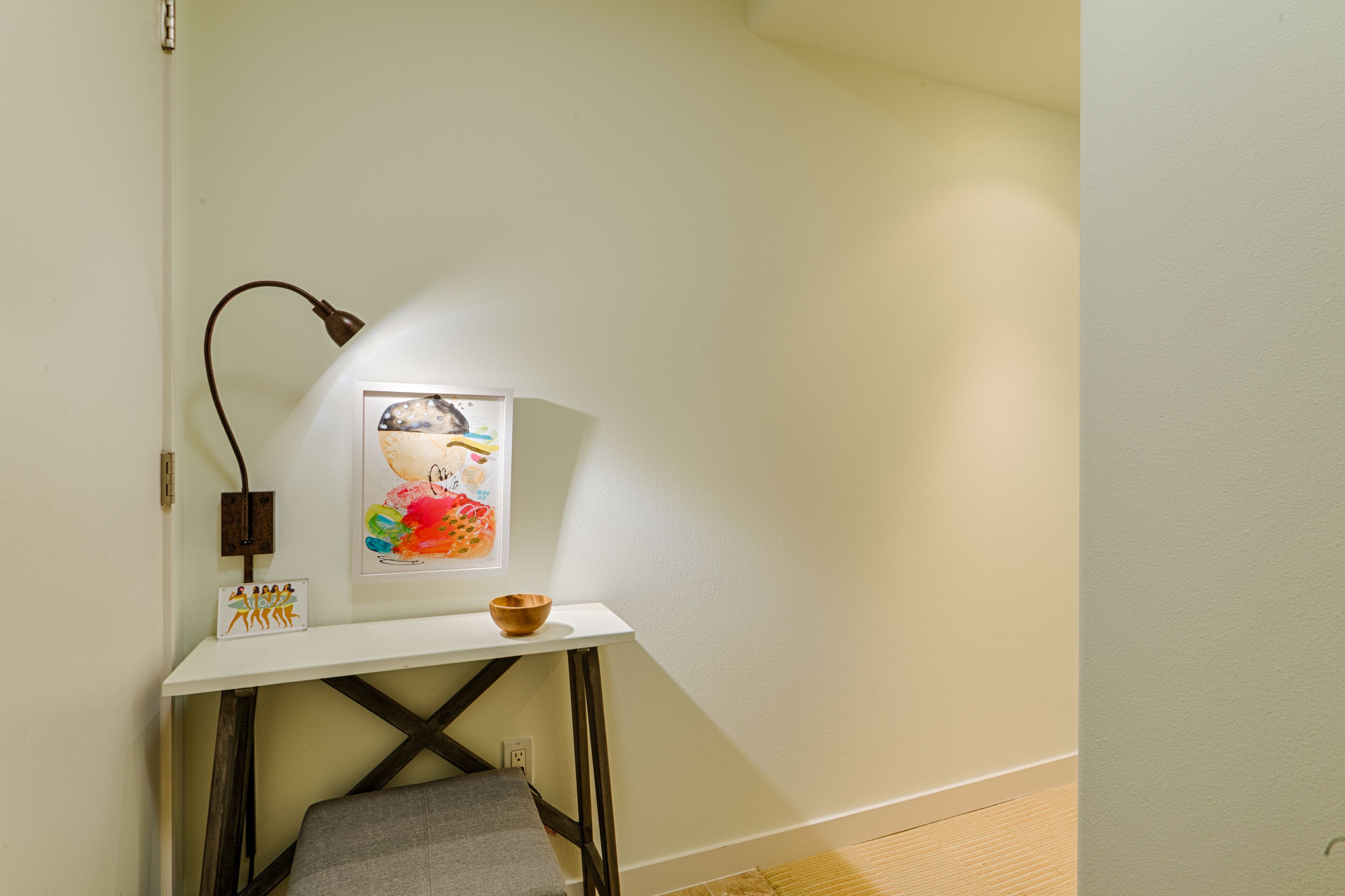 A corner room with a white wall, a small white table, a lamp, a framed picture, and a wooden bowl.