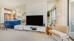 Un salon moderne doté d’un grand téléviseur à écran plat, d’un meuble de rangement blanc, d’un canapé et d’un coin repas avec des tabourets de bar.