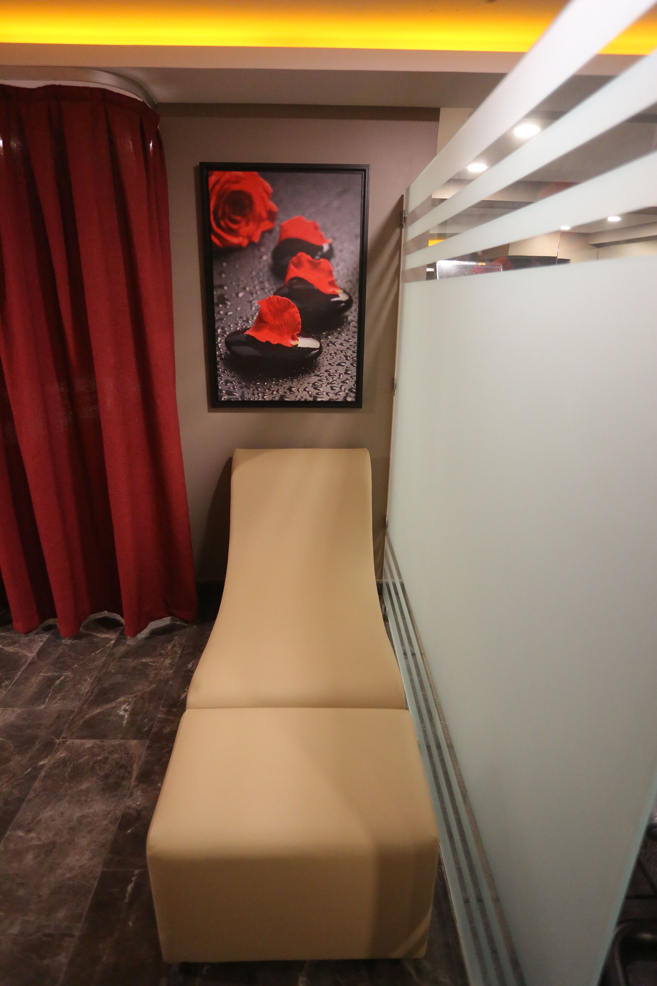 Une pièce avec une table de massage beige, des rideaux rouges et un tableau représentant des roses rouges accroché au mur.