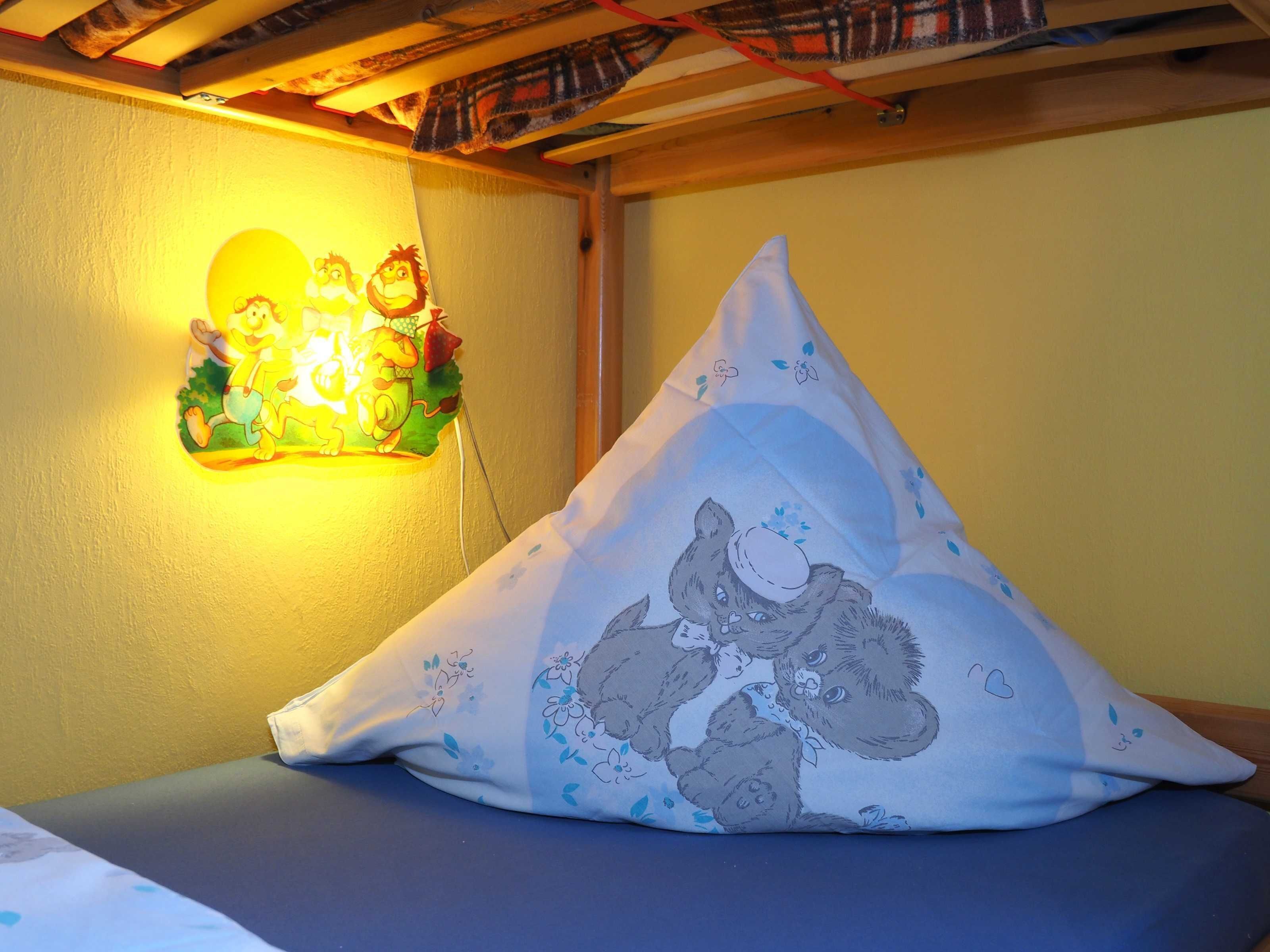 Una cama con un almohadón de personajes de dibujos animados, una lámpara de pared con una escena caricaturesca y un techo de madera adornado con tela estampada.