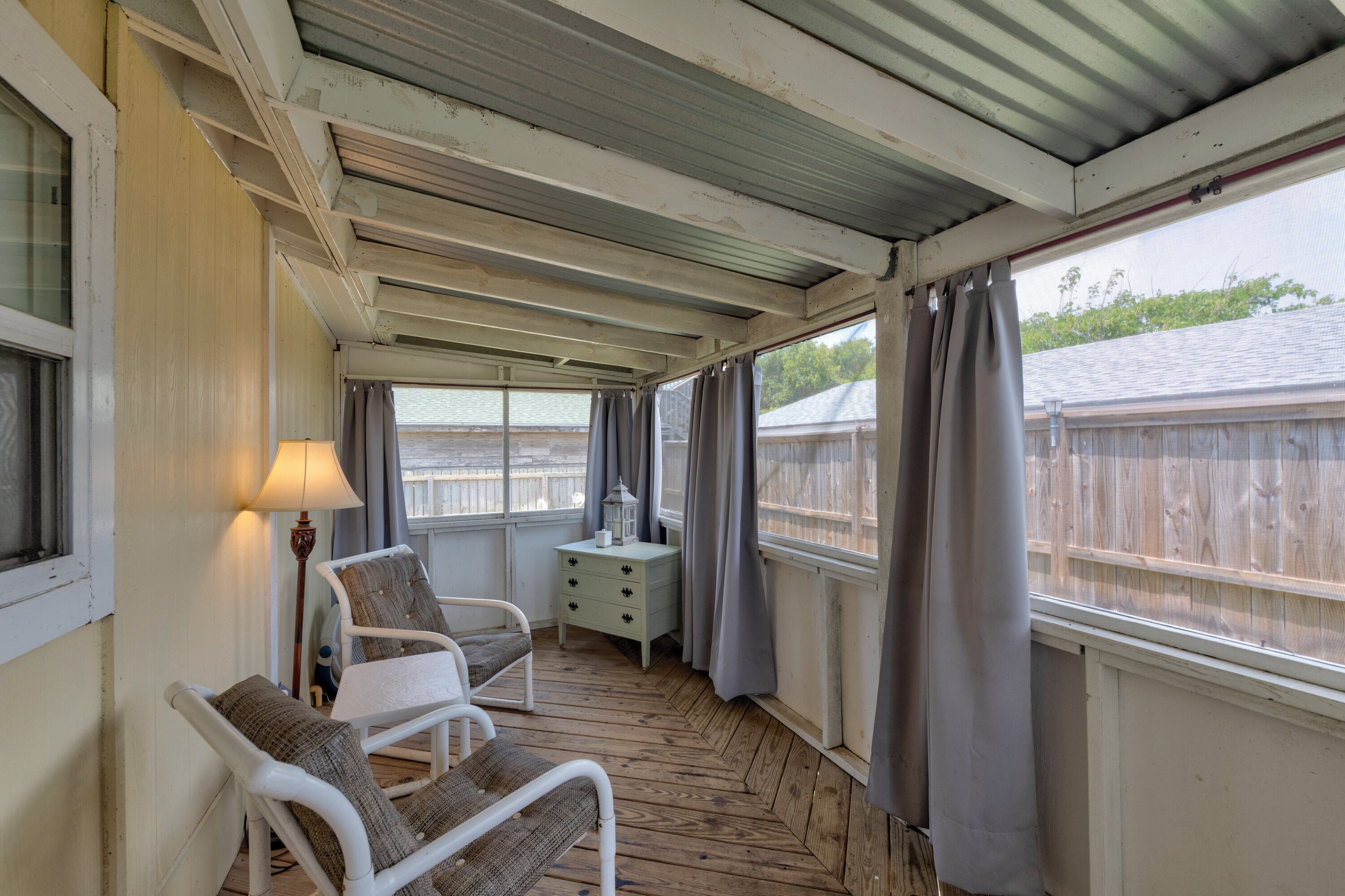 A covered balcony with wooden flooring, a white chair, a small table, a lamp, and a white cabinet.