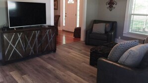 A living room with a flat-screen TV on a wooden cabinet, a dark leather sofa, and a patterned ottoman. - Back to School Special! 5 minutes from Majestic Baseball Fields (Hot Springs)