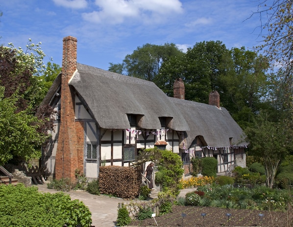 A traditional half-timbered house with a thatched roof, brick chimneys, and a garden with various plants and flowers.