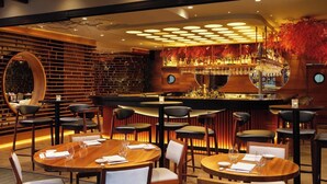 Un restaurant moderne avec des tables et des chaises rondes, un coin bar agrémenté d’un grand miroir circulaire et un intérieur chaleureux au décor en bois.