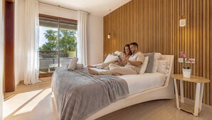 Een moderne slaapkamer met een gebogen bed, een klein nachtkastje met een potplant, en een uitzicht op palmbomen door een groot raam.