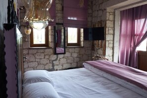 A bedroom with a stone wall, a bed with white bedding, a television mounted on the wall, and a window with a view of the outside. - Sila Alacati (Cesme)