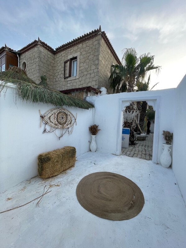A courtyard with a stone building, a white wall, a woven mat, a hay bale, and a decorative wall hanging. - Sila Alacati (Cesme)