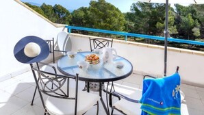 Ein runder Glastisch mit Stühlen, einem weißen Sonnenschirm und einem blauen Handtuch auf einem Balkon mit Geländer und Bäumen im Hintergrund.