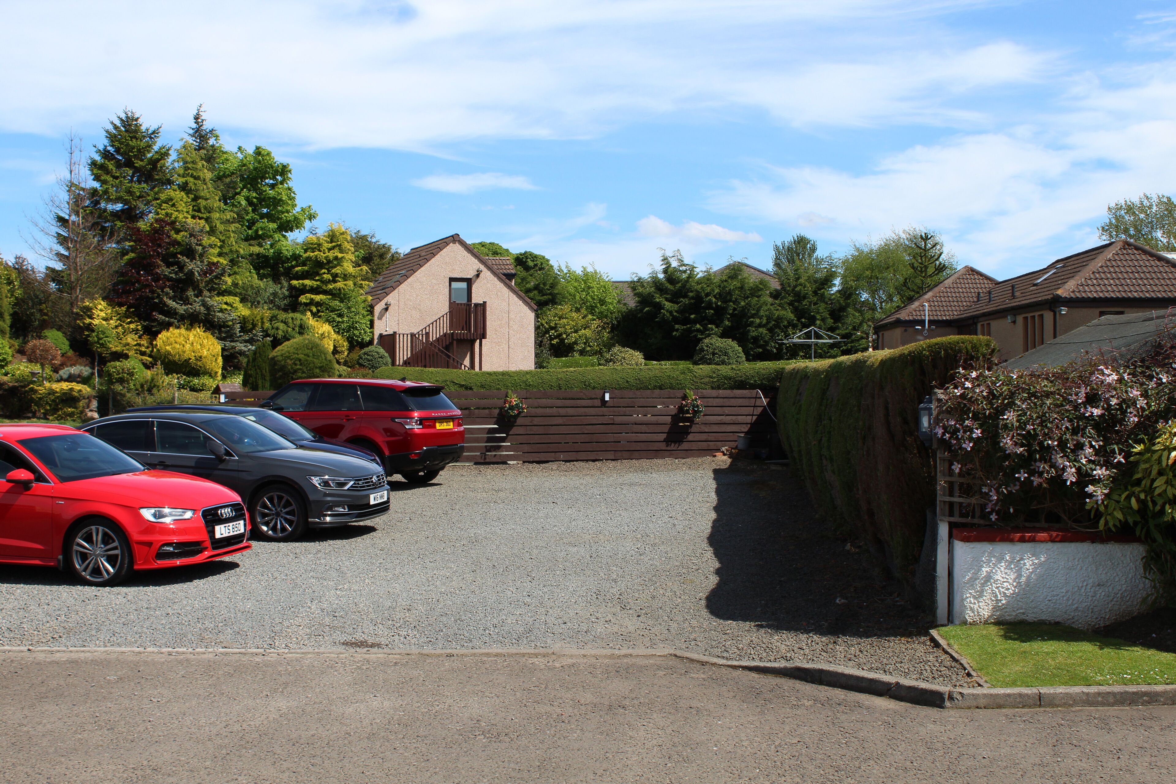 A parking area with several cars, a building with a sloped roof, and a well-maintained hedge.