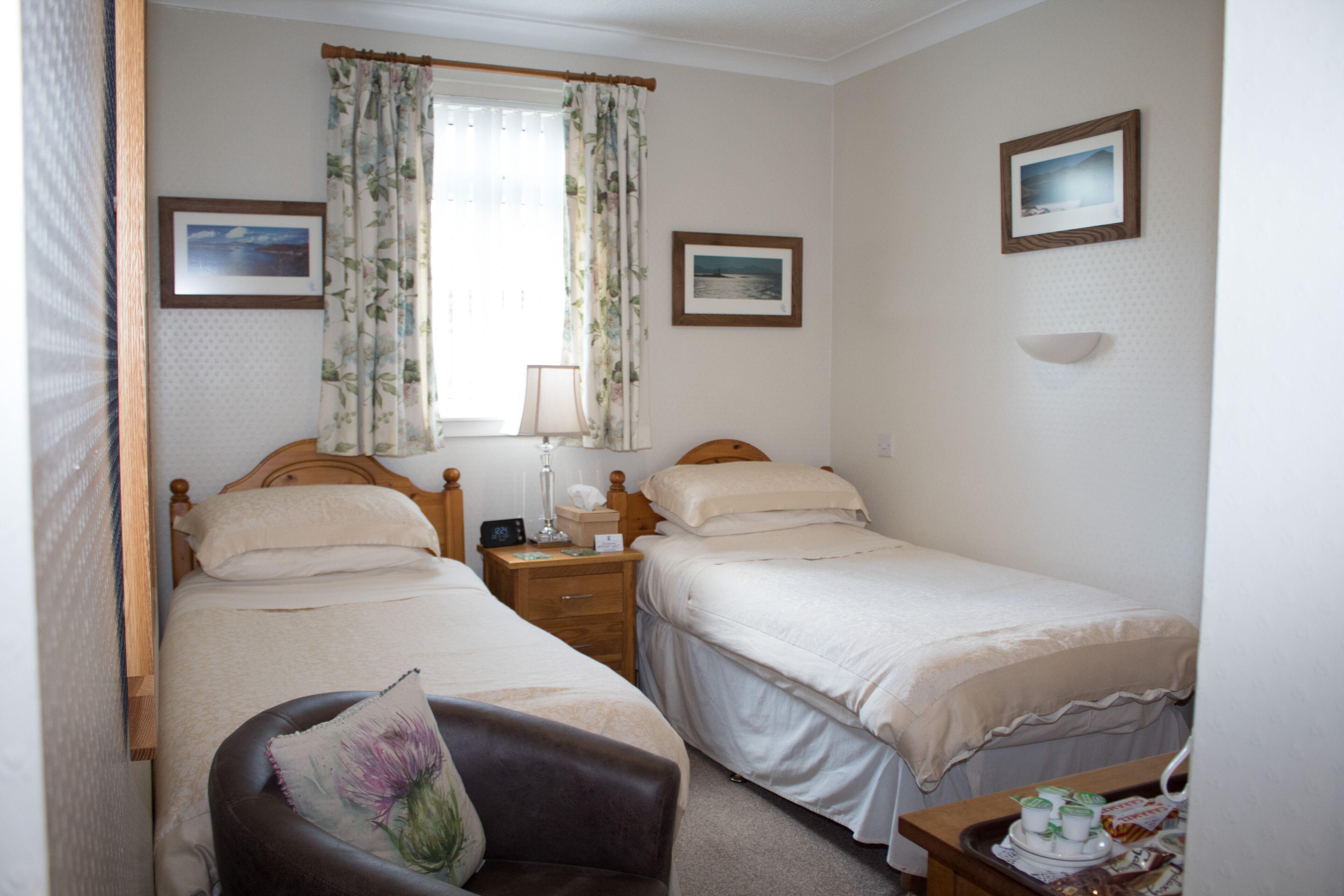 Une chambre avec deux lits simples, une table de chevet avec une lampe, une chaise et une petite table sur laquelle se trouve un plateau avec divers objets.