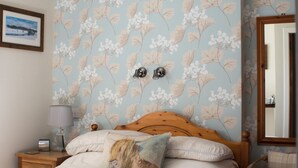 Ein Schlafzimmer mit floraler Tapete, einem hölzernen Bett, einem Nachttisch mit Lampe, einem Sessel und einem Spiegel.