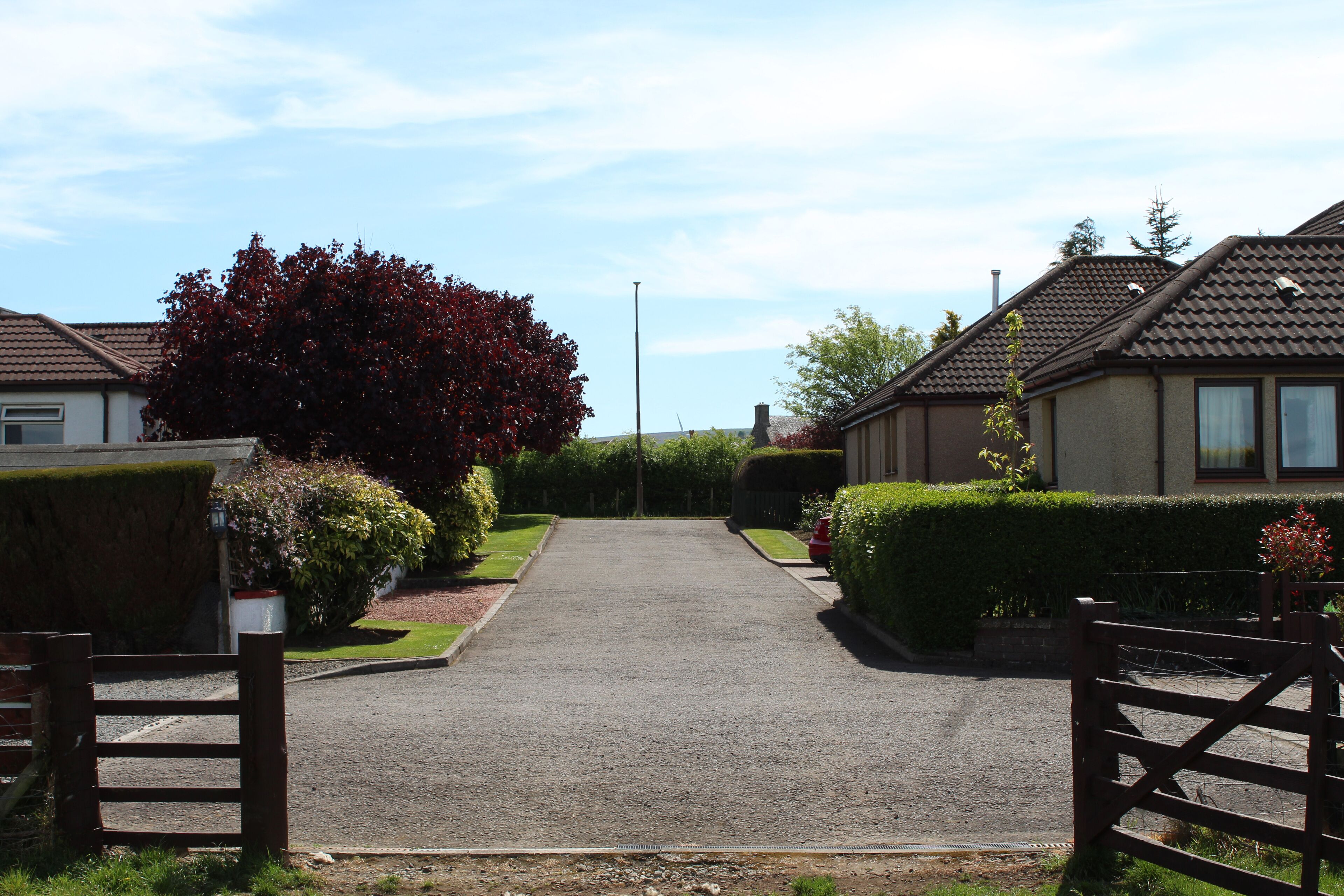 A residential street with houses on either side, a gravel driveway, and a wooden gate at the entrance.