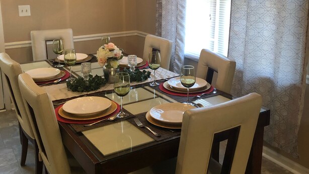 Comedor con una mesa rectangular dispuesta para seis personas, rodeada por seis sillas. Un candelabro cuelga del techo, y se ve una ventana con cortinas estampadas.