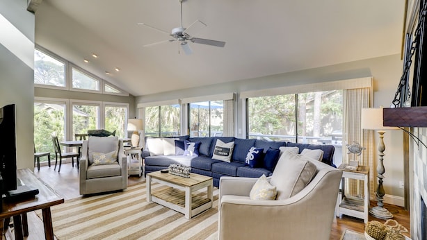 A spacious living room with a large sofa, a coffee table, and a television. There are large windows providing natural light and views of the outdoors. - Four Bedroom Sea Pines Home with Private Pool ~ 5 Minute Walk to the Beach! (Hilton Head Island)
