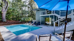 A modern outdoor pool area with a blue umbrella, lounge chairs, and a staircase leading to a second-floor deck. - Four Bedroom Sea Pines Home with Private Pool ~ 5 Minute Walk to the Beach! (Hilton Head Island)