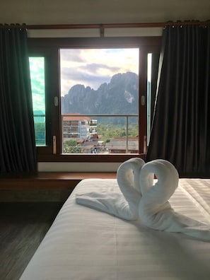 A hotel room with a bed, a window with curtains, and a view of mountains and buildings. - Faraway Suites - Hostel (Vang Vieng)