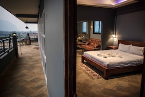 A bedroom with a large bed, a wooden headboard, a nightstand, a chair, and a small table. There is a balcony with a railing, a view of the mountains, and a table with a lantern. - Faraway Suites - Hostel (Vang Vieng)
