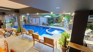 A pool area with wooden chairs and tables, surrounded by potted plants and a view of a garden. - Faraway Suites - Hostel (Vang Vieng)