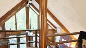 Slaapkamer in een loft met schuine plafonds, houten balken, uitzicht op de omgeving, een plafondventilator en een raam met gordijnen.