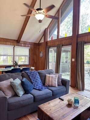 A cozy living room with a gray sofa, wooden ceiling beams, a ceiling fan, and large windows with curtains. - Bedrooms 3, Bathrooms 1, Sleeps 6 (Linden)