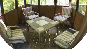 Een overdekt terras met houten terras, een tafel met stoelen en grote ramen met uitzicht op de omliggende bomen.