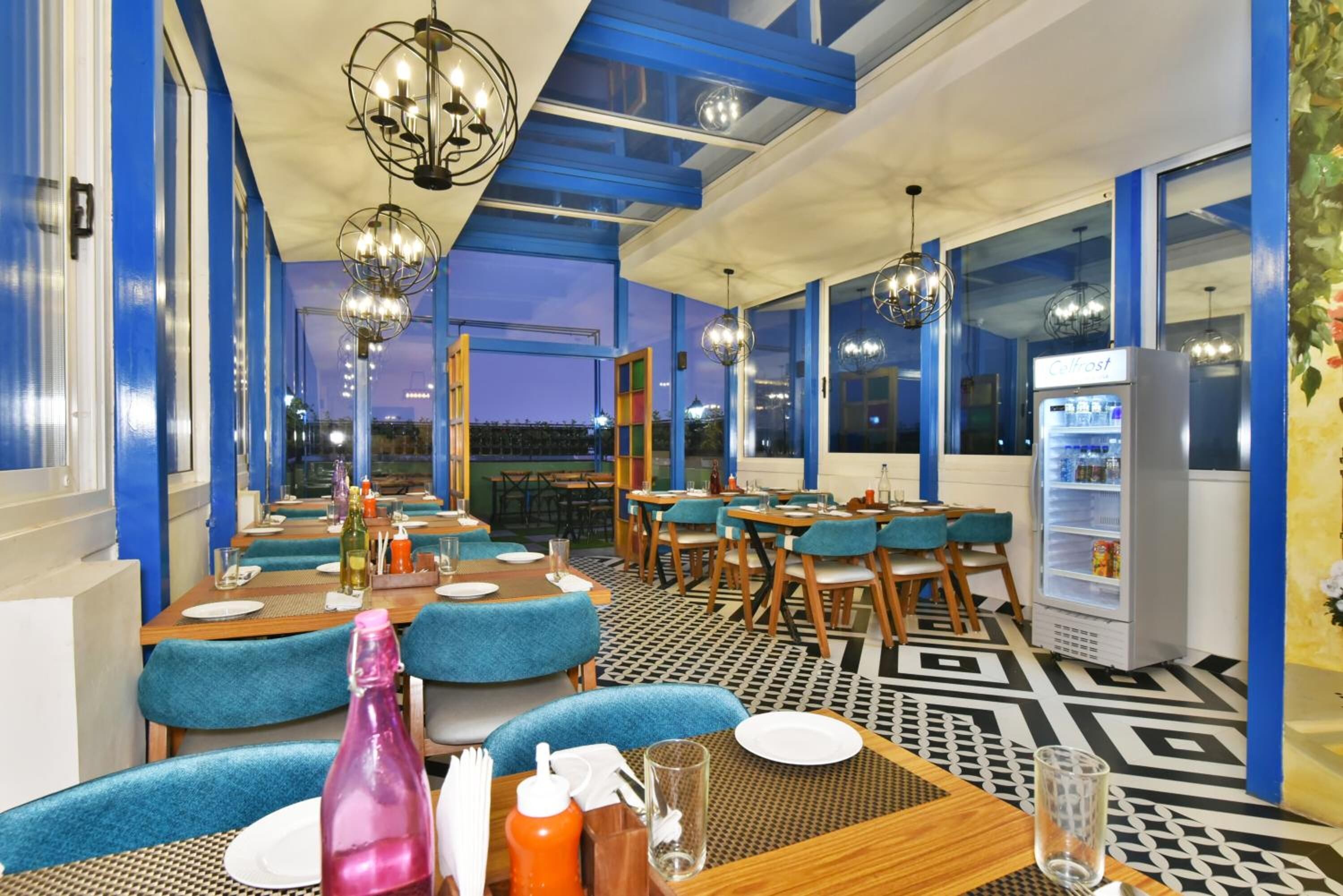 Un restaurante con paredes azules, mesas y sillas de madera. Hay luces colgantes y un refrigerador en la pared.