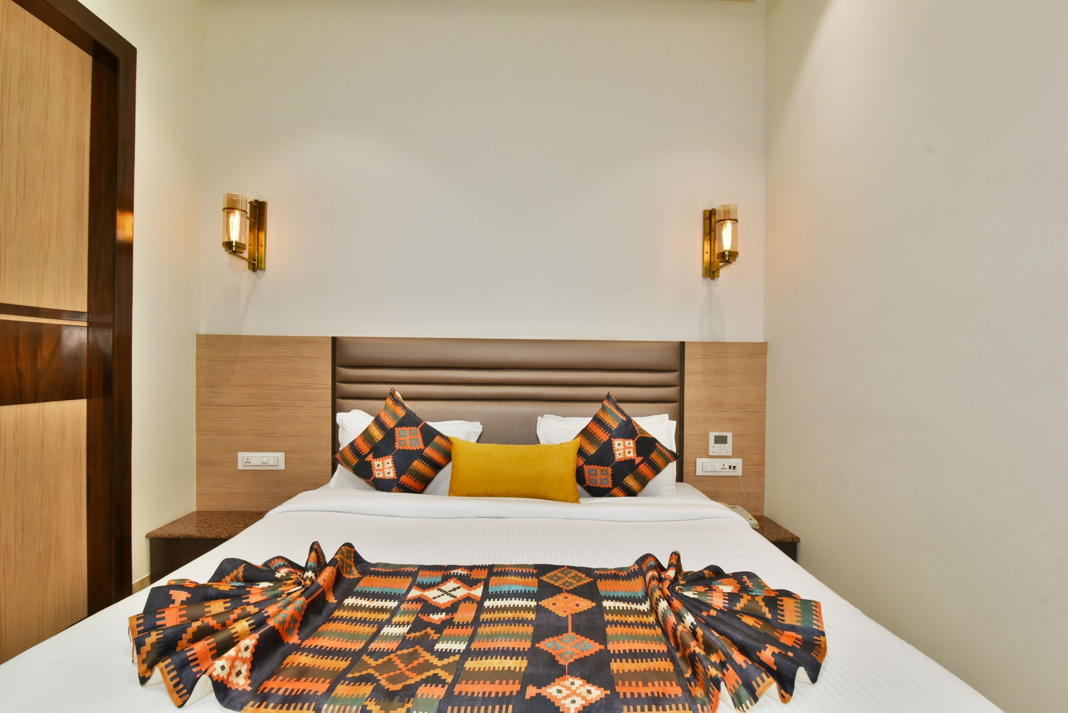 A hotel room with a bed, two wall-mounted light fixtures, a wooden headboard, and a patterned blanket.