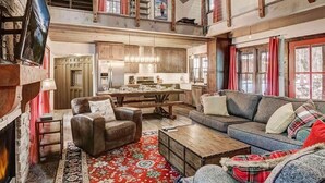 A spacious living area with a fireplace, a sofa, a coffee table, and a dining table. There is a kitchen in the background with wooden cabinets and a window. - Heavenly Hideaway 4BR,4BA, Hot Tub (South Lake Tahoe)