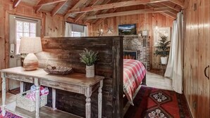 A rustic cabin interior with exposed wooden beams, a fireplace, a bed with a red and white checkered blanket, and a table with a lamp and decorative items. - Heavenly Hideaway 4BR,4BA, Hot Tub (South Lake Tahoe)