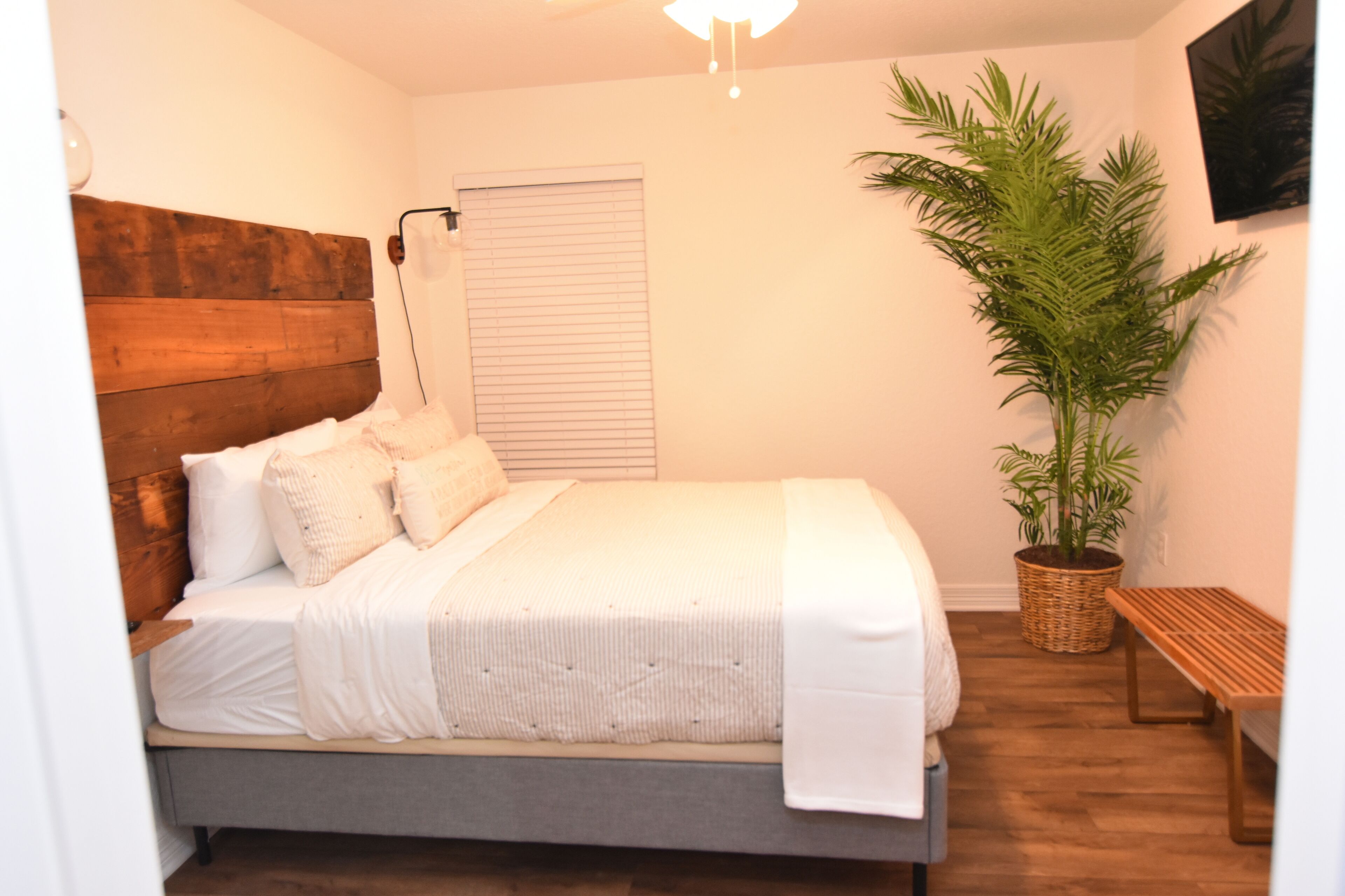 A bedroom with a wooden headboard, a large potted plant, a bench, and a window with blinds.