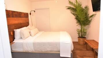 A bedroom with a wooden headboard, a large potted plant, a bench, and a window with blinds.