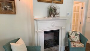 Ein Wohnzimmer mit einem weißen Kamin, zwei smaragdgrünen Sesseln mit floralen Kissen und einem Stapel Bücher auf einem kleinen Tisch.