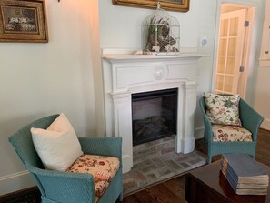 Een woonkamer met een witte open haard, twee blauwgroene fauteuils met bloemenkussens, en een stapel boeken op een bijzettafeltje.