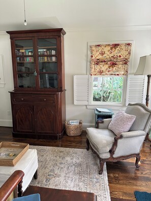 A room with a wooden bookcase, a patterned window treatment, a chair with a cushion, and a wicker basket.