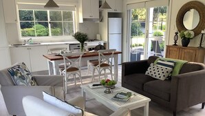Una sala moderna con un sofá blanco, un sofá marrón y una mesa de centro con un bol de manzanas. Hay un comedor con sillas blancas y una mesa de madera.