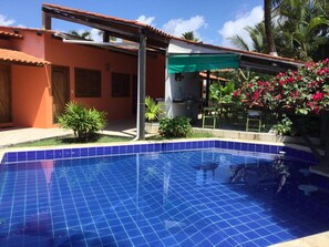 A swimming pool with blue tiles, surrounded by a patio area with a covered seating area and potted plants.