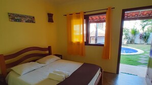 A bedroom with a wooden bed, a window with curtains, and a view of a garden. - Pousada Villa Tropicale (Salvador)