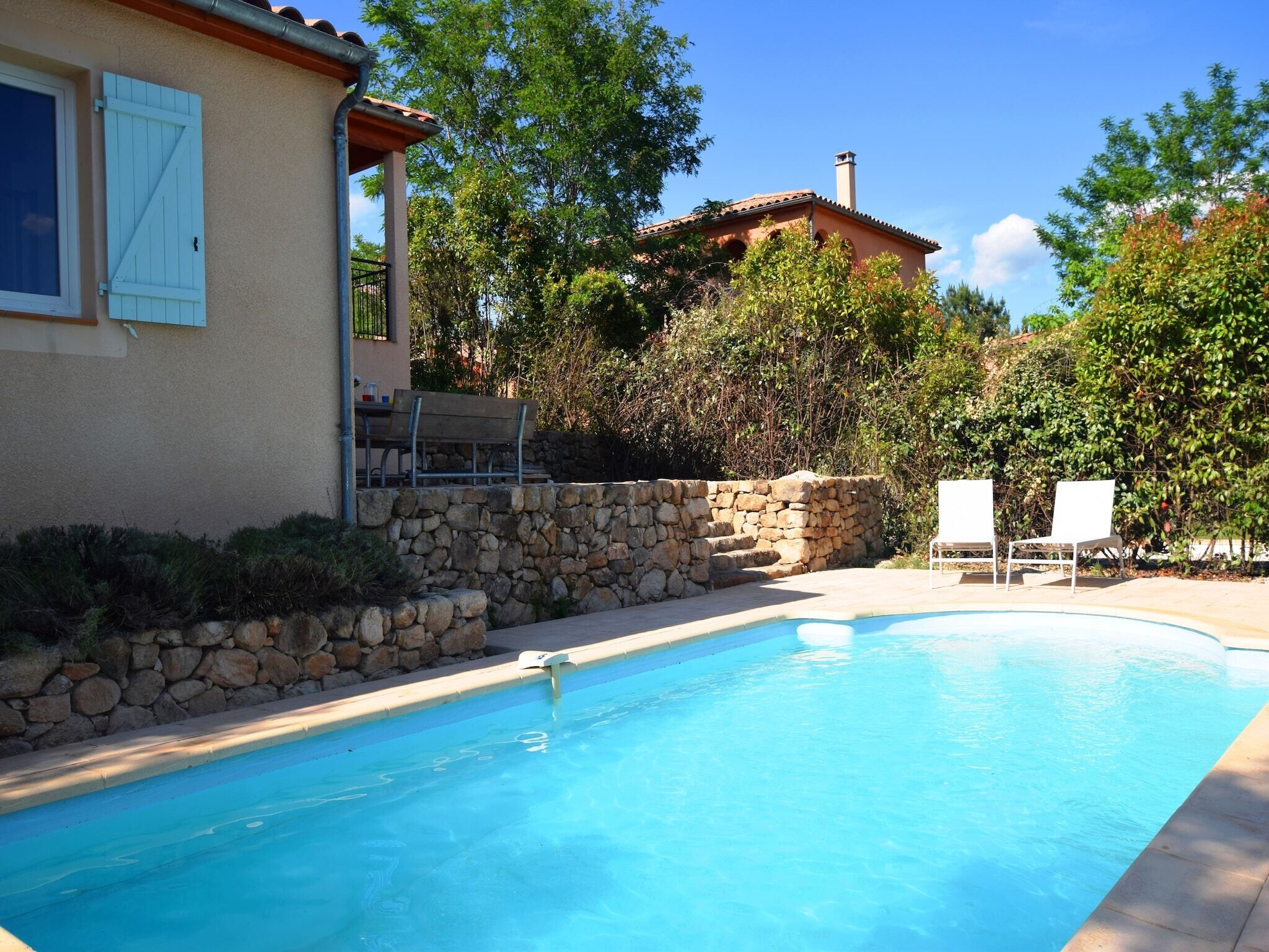 A swimming pool with stone walls and a bench, surrounded by greenery and a house with a tiled roof.