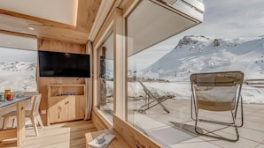 Een gezellige kamer met houten inrichting, een flatscreen televisie, een eethoek en een groot raam met uitzicht op een besneeuwd berglandschap.
