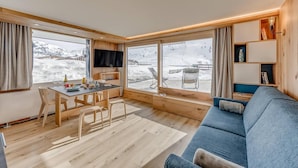Een gezellige kamer met houten vloer, een blauwe bank, een eettafel met stoelen en grote ramen met uitzicht op een besneeuwde berg.