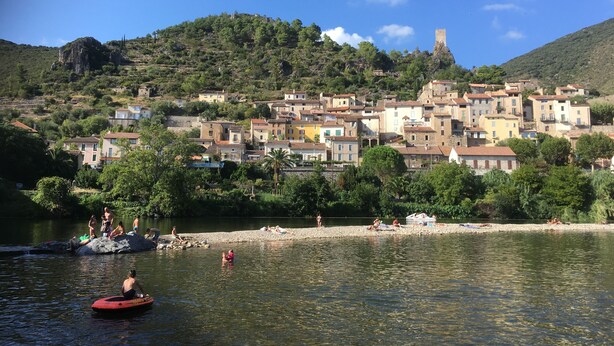 Un pueblo costero con edificios en una ladera, un río en primer plano y gente disfrutando de actividades acuáticas.