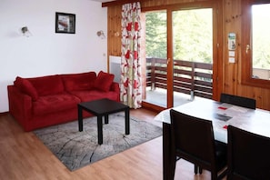 A room with a red sofa, a small black table, a dining table with chairs, a wooden deck, and a sliding glass door.
