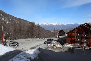 A mountain lodge with a view of snow-capped peaks, a parking area with several cars, and a wooden building with balconies.