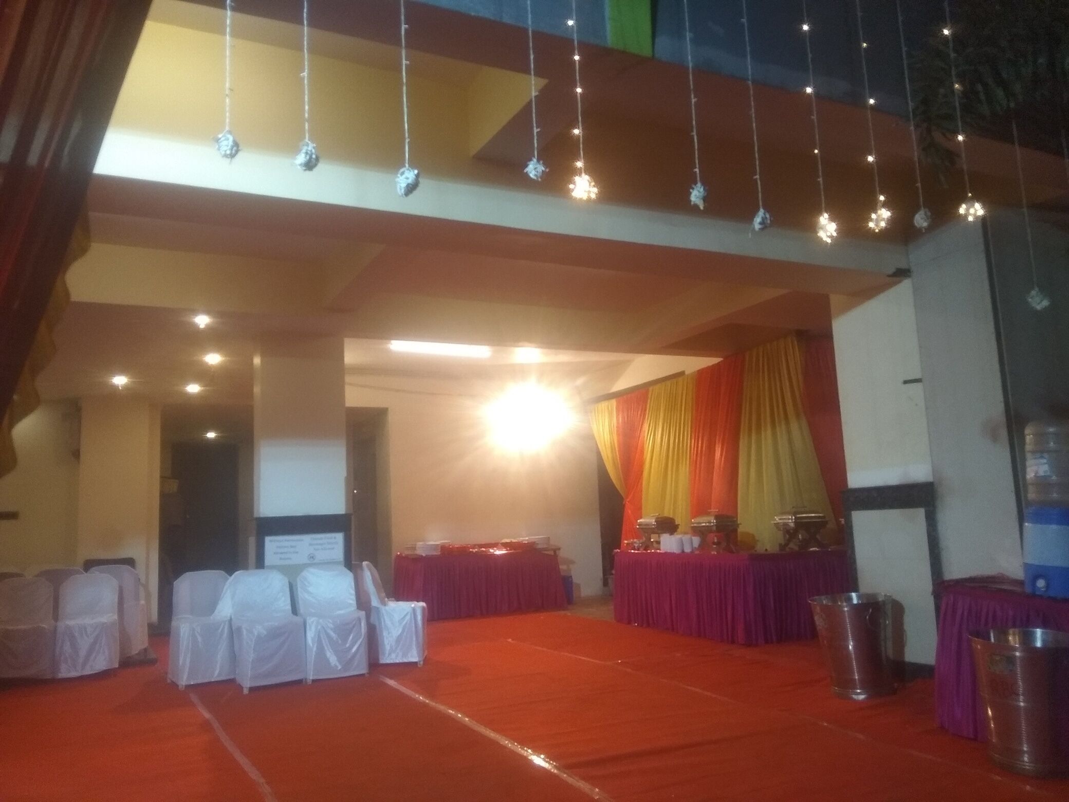 Un salón de banquetes con filas de sillas cubiertas de blanco, mesas con manteles morados y luces colgantes.