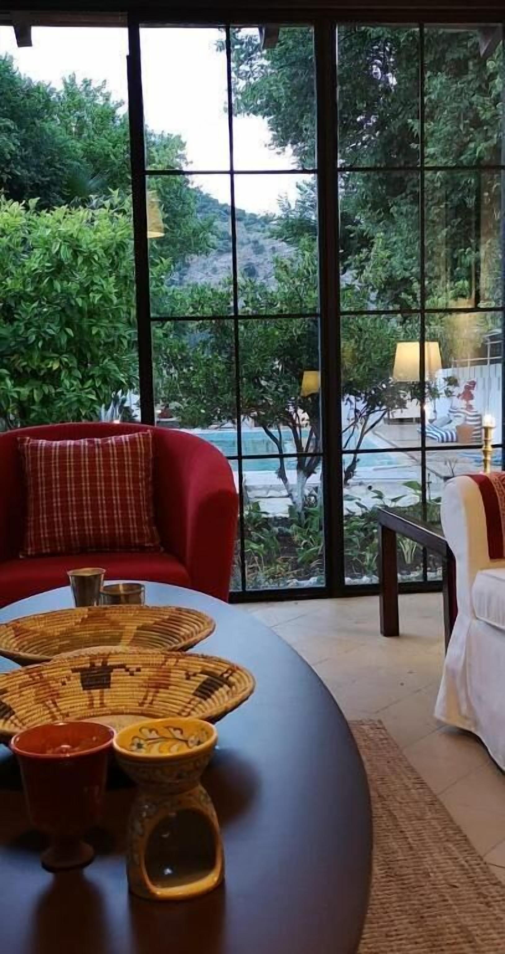 Ein Zimmer mit großem Fenster, einem roten Sofa, einem Tisch mit gewebten Untersetzern und Blick auf einen Pool und Grünflächen.