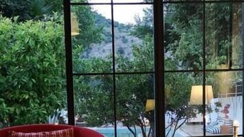 Una habitación con un ventanal grande, un sofá rojo, una mesa con individuales tejidos y vistas a una piscina y follaje.
