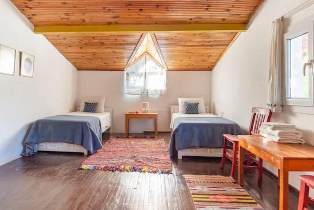 Una habitación con dos camas, techo de madera, una ventana y una pequeña mesa con una silla.