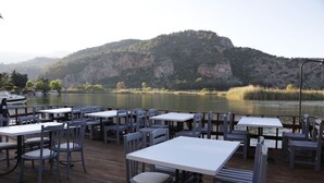 An outdoor dining area with tables and chairs, overlooking a lake with boats and a mountainous backdrop. - Dalyan Holiday Hotel (Ortaca)